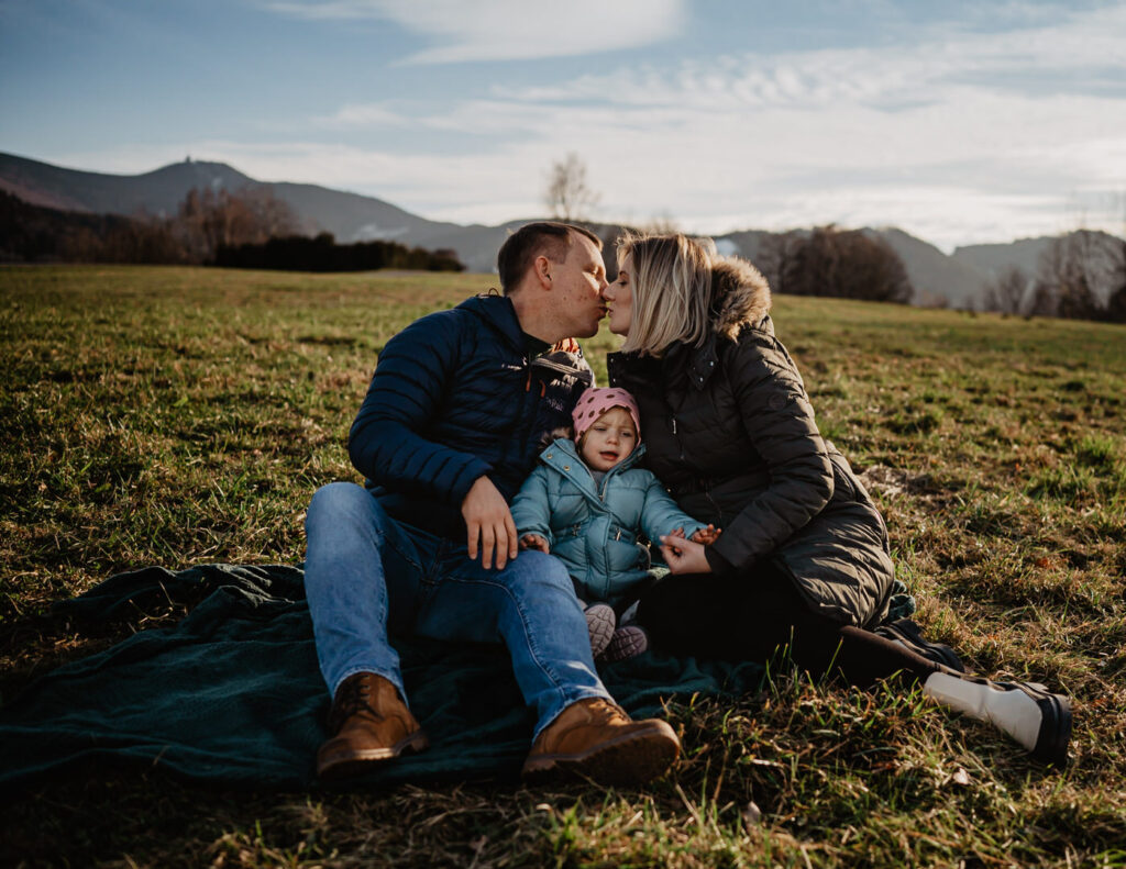 a man and woman kissing a child on a blanket in a grassy field